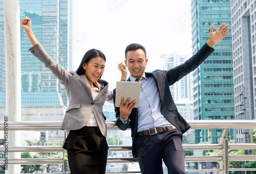 Young happy Asian businessman and budinesswoman looking in tablet standing in front of office buildings caught in the moment of joy for the successful completion of the job. photo