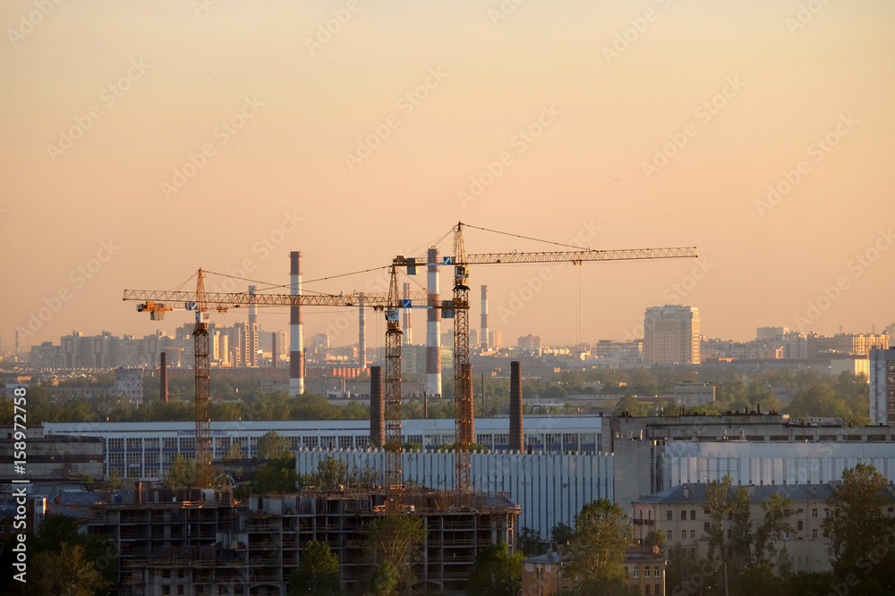 Photo of a building process in the city in the sunset. Cranes and pipes.