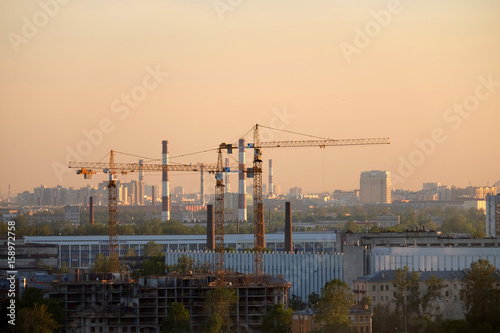 Photo of a building process in the city in the sunset. Cranes and pipes.