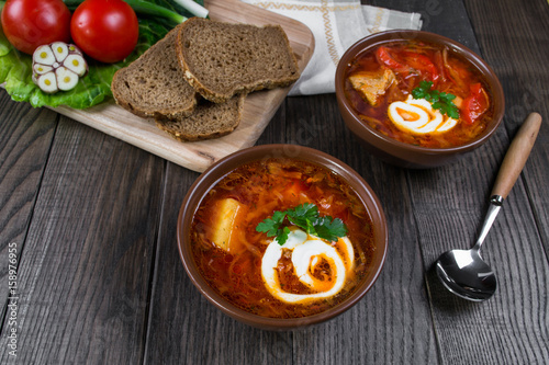 Tomato soup . Traditional  Ukrainian beetroot and tomato soup - borsch in clay pot with sour cream, garlic, herbs and bread on dark wooden background.Ingredients on table