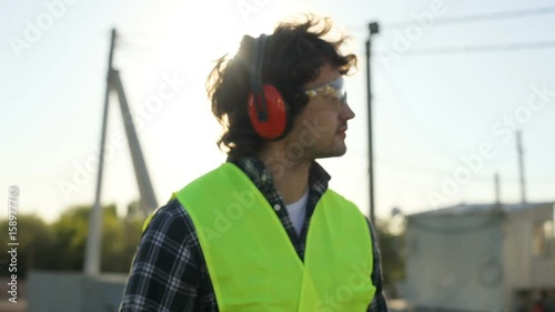 Side view of young caucasian builder with glasses and headphones in a green vest going at initial stage of construction. Outdoor. photo