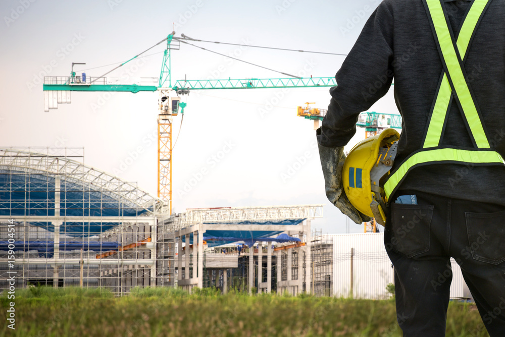 Rear view Asian male site contractor engineer with hard hat holding blue print paper inspecting at construction site, crane with golden sunlight at the background. Rear view Asian male site contractor engineer with hard hat holding blue print paper inspecting at construction site, crane with golden sunlight at the background.