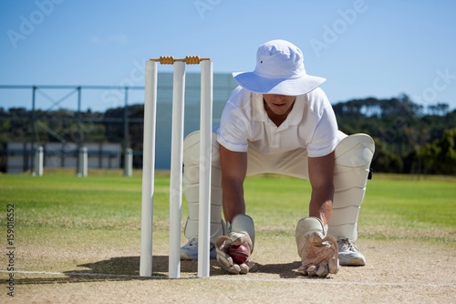 Wicketkeeper holding ball behind stumps against blue sky