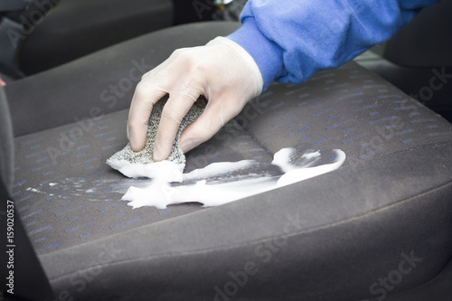 Ręka w rękawicy ochronnej pierze tapicerkę fotela samochodowego za pomocą pianki. Ręczne pranie tapicerki samochodowej za pomocą pianki do prania i gąbki.