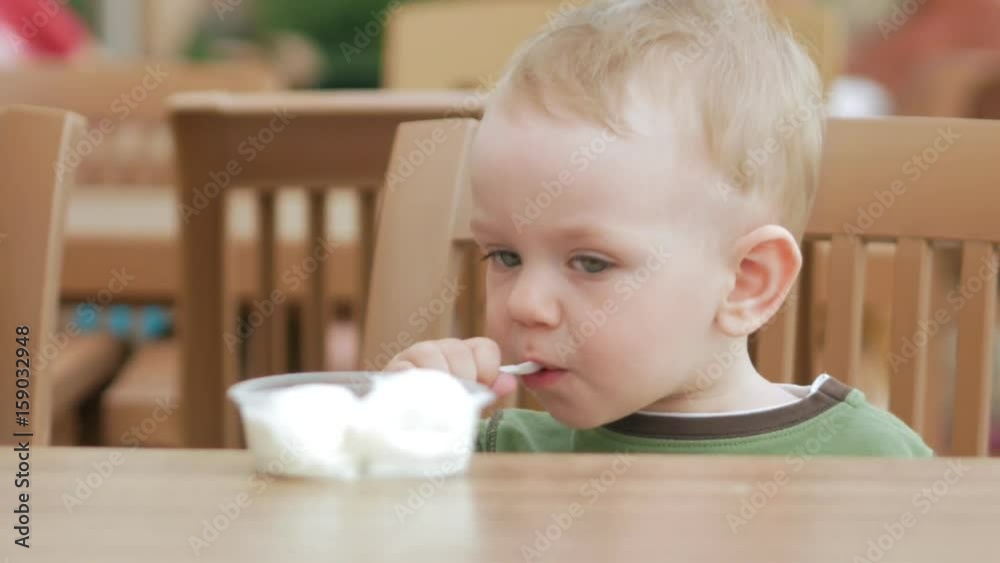 Mother feeds her young son with ice cream from a spoon. They are sitting in a street cafe. The boy really likes the delicacy. Mother feeds her young son with ice cream from a spoon. They are sitting in a street cafe. The boy really likes the delicacy.