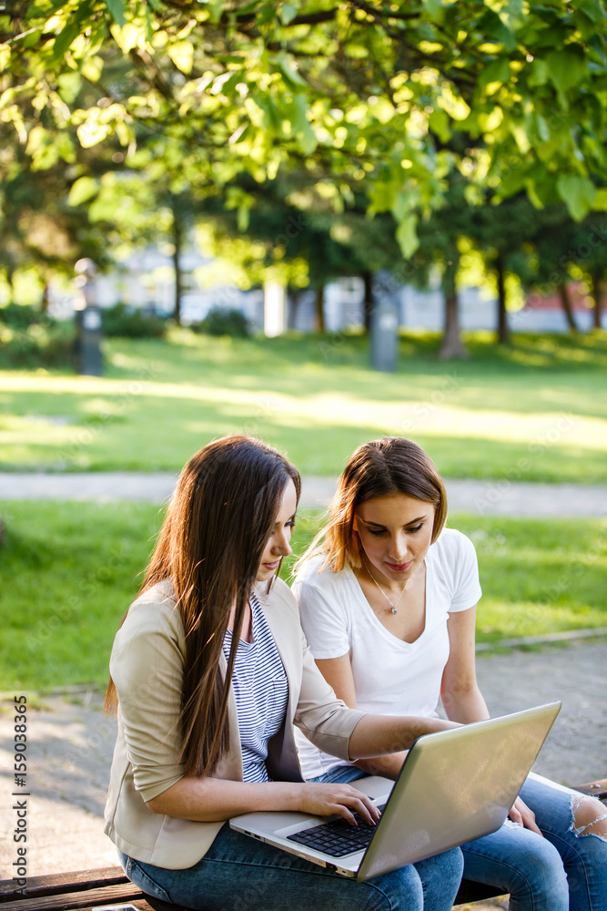 Two female friends in park sitting on bench and working on laptop