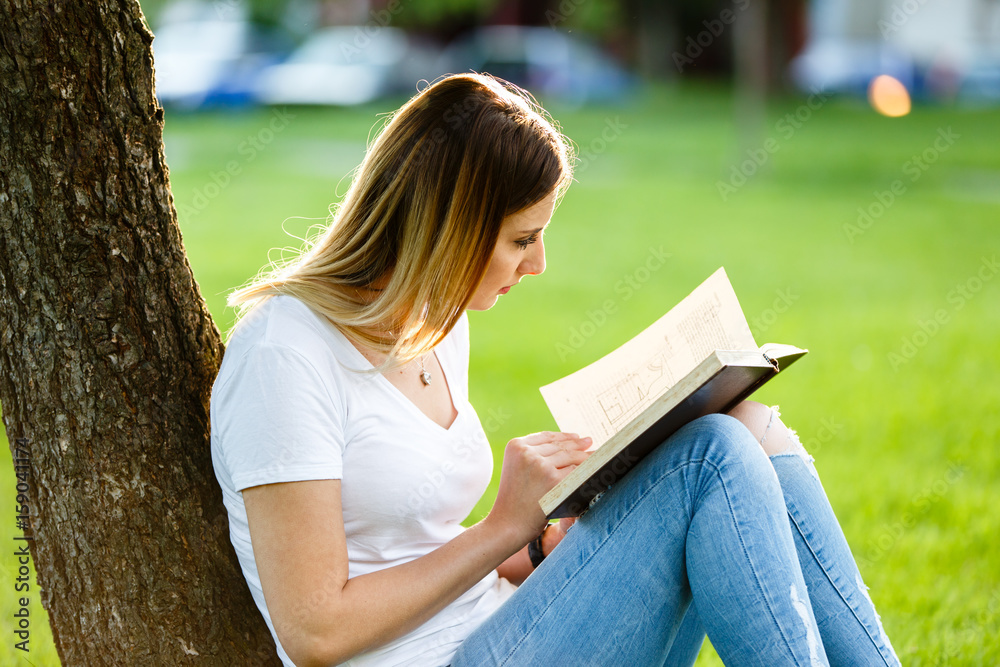 Obraz premium Young girl sitting in park and reading a book under the tree