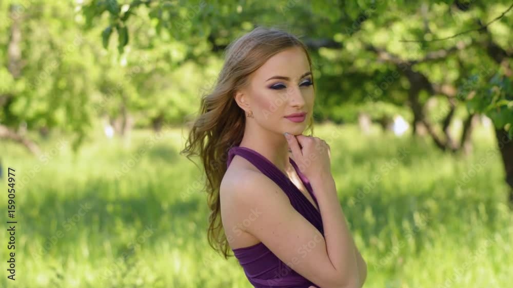 Portrait of a beautiful girl with bright make-up. Caucasian blonde woman posing and looking at camera in lilac or purple dress in green garden near apple fruit tree. Portrait of a beautiful girl with bright make-up. Caucasian blonde woman posing and looking at camera in lilac or purple dress in green garden near apple fruit tree.