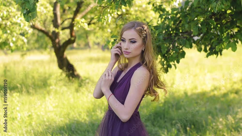 Portrait of a beautiful girl with bright make-up. Caucasian blonde woman posing and looking at camera in lilac or purple dress in green garden near apple fruit tree. Portrait of a beautiful girl with bright make-up. Caucasian blonde woman posing and looking at camera in lilac or purple dress in green garden near apple fruit tree.