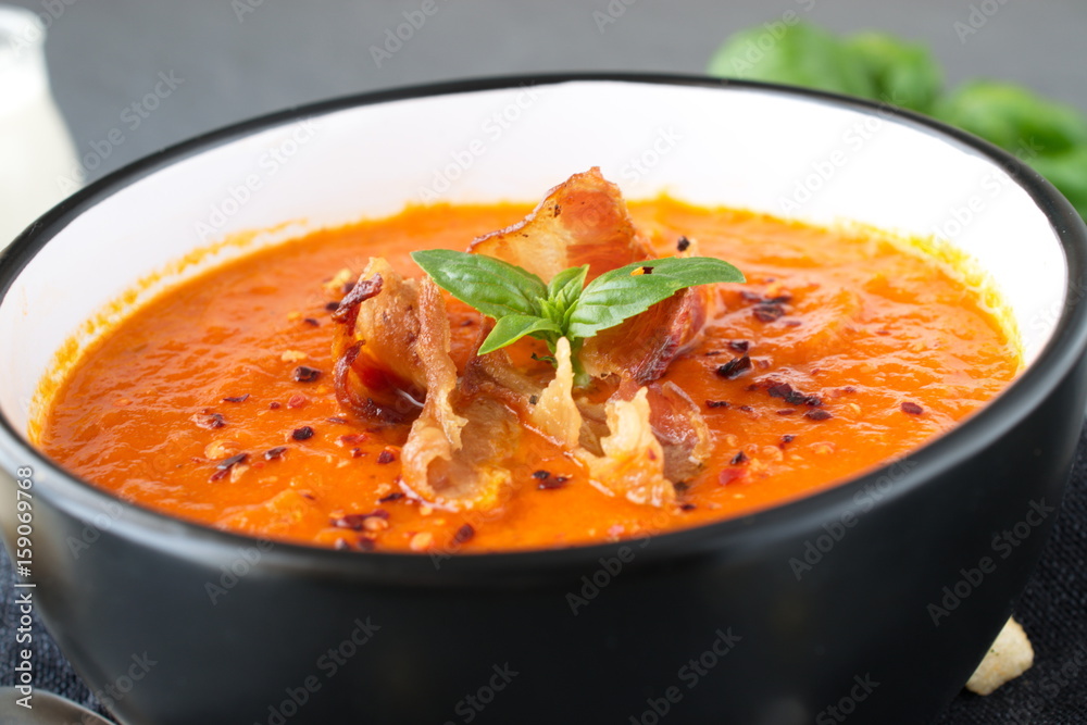Thick tomato soup with basil and fried bacon in a black ceramic bowl on a grey abstract background. Healthy eating concept Fresh tomato soup with creme fraiche Thick tomato soup with basil and fried bacon in a black ceramic bowl on a grey abstract background. Healthy eating concept Fresh tomato soup with creme fraiche