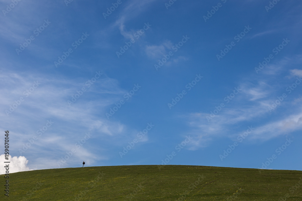 A small human figure on top of a green hill beneath a huge sky