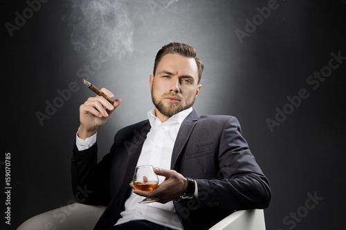 Portrait of handsome young man with cigar and cognac glass. Perfect hair style, beard and suit. Studio grey background