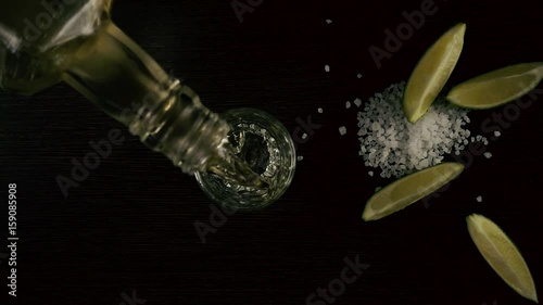 Slow motion. The bartender from the bottle will pour the tequila in a glass on the table and next to the lime and salt. Top view