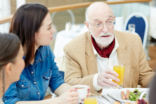 Man with grey beard holding glass of orange juice while talking to his family by breakfast
