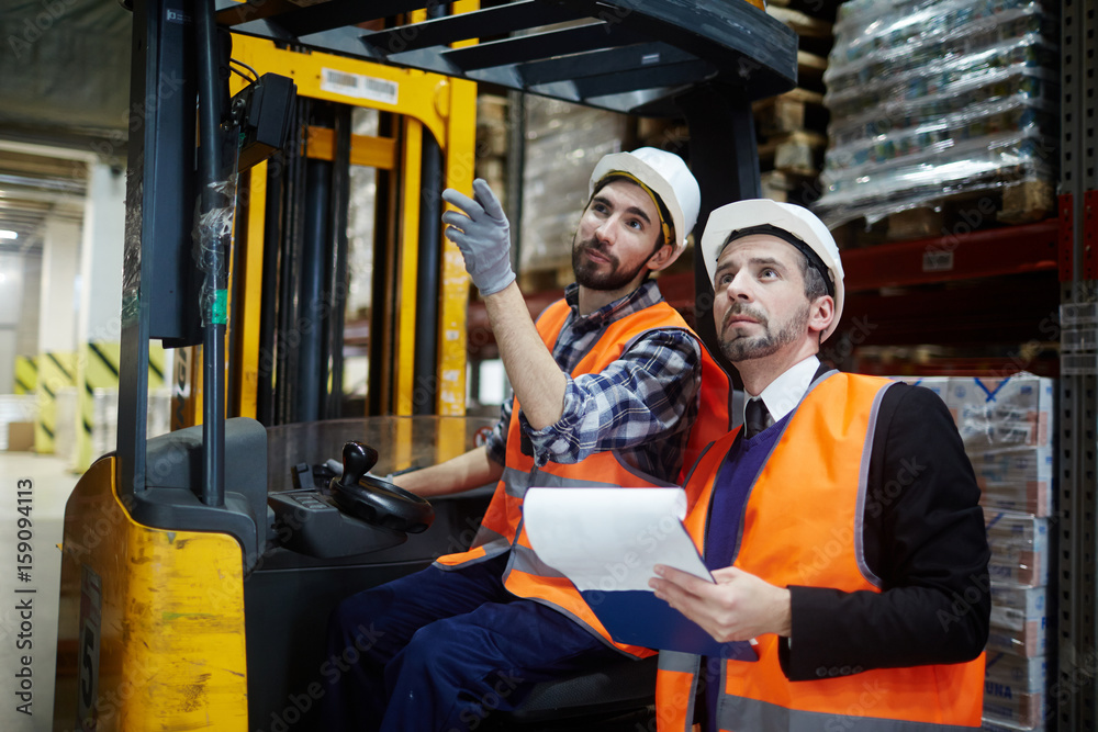 Forklift worker pointing at one of warehous shelves while talking to his master