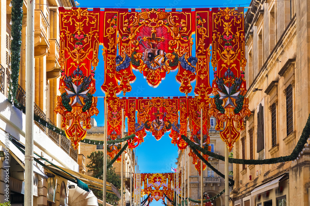 Naklejka premium Festively decorated street with banners for St Augustine Feast in the old town of Valletta, Malta. Flaming, arrow pierced heart - symbol of St Augustine