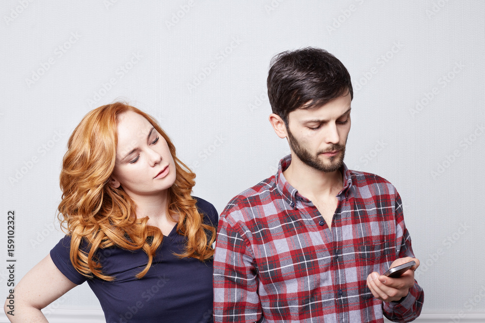 Handsome Caucasian male with beard reading sms on mobile phone, messaging lover while his possessive beautiful wife spying on him, looking at screen of electronic gadget with sneaky expression Handsome Caucasian male with beard reading sms on mobile phone, messaging lover while his possessive beautiful wife spying on him, looking at screen of electronic gadget with sneaky expression