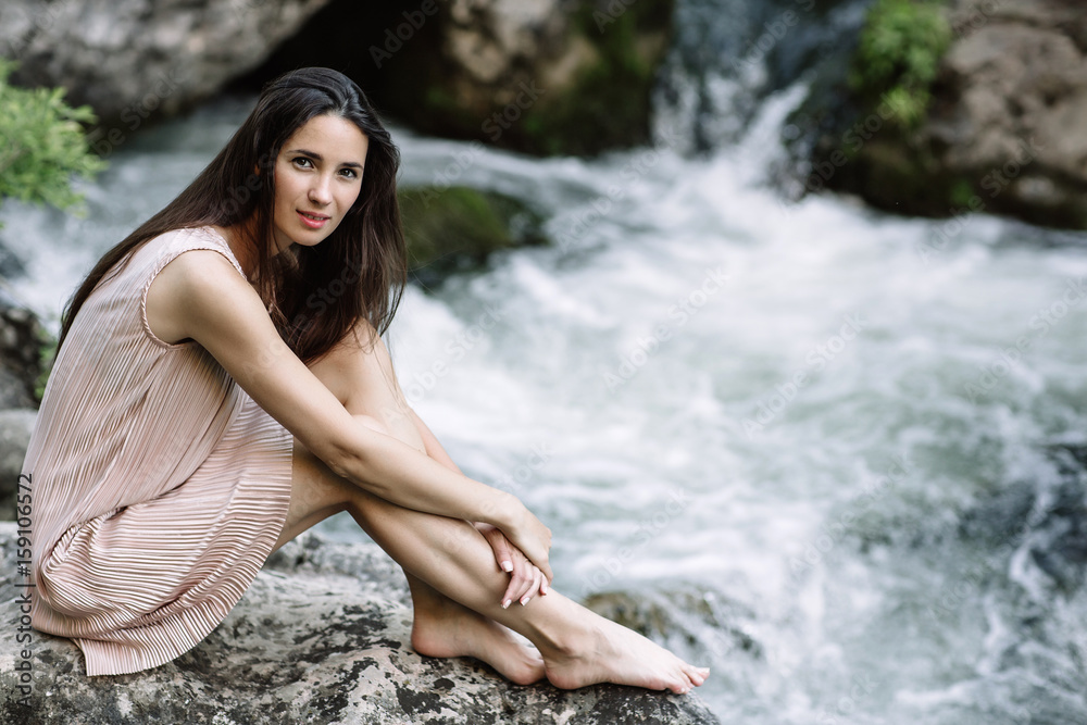 Beautiful brunette sitting on a rock in the middle of the river, water, waterfall. Beautiful nature, natural light, soft, filling