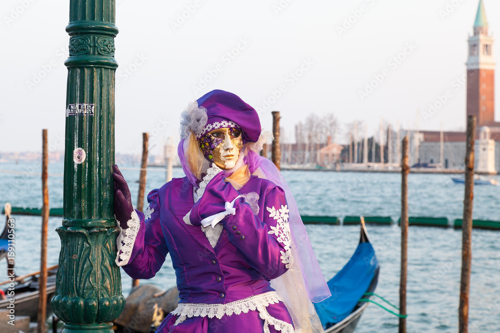 2017 Venice  Carnival, Veneto, Italy. Woman in classical purple mask and costume posing at sunset  in front of gondolas at the lagoon at Piazza San Marco  with San Giorgio Maggiore behind 
