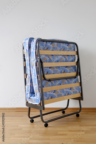 A folding bed in front of a white wall an a blue mattress.