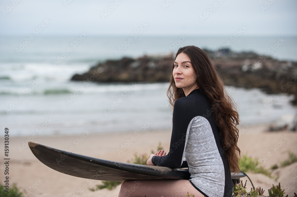 Sexy California surfer girl holding surf board across lap smiling to left. 