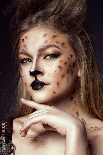Close up portrait of young beautiful grey-eyed model with artistic leopard make-up and brushed up hair looking straight at the camera with her finger under the chin. Studio shot