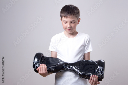 A teenager in a white T-shirt is holding a black self-balancing two-wheeled hoverboard and looks at him. On a gray background. photo