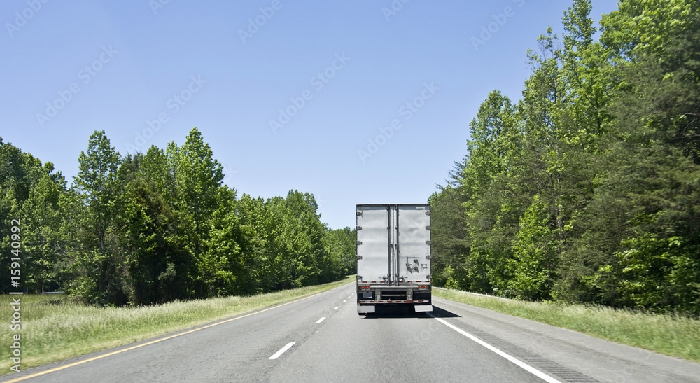 Rear view of semi truck and trailer on highway flanked by trees with blue sky.