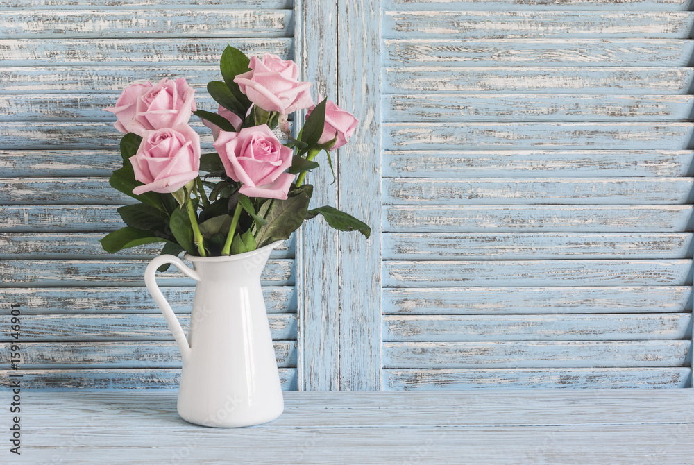 Obraz premium Pink roses in white enamel jug on a blue rustic background. Free space for text. Vintage style