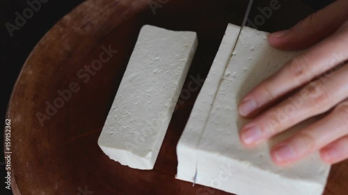 Close up of female hands cutting tofu (made of soy beans) in big slices on round wooden board. Tofu is mainly used in Asian and vegetarian cooking. Table top view.