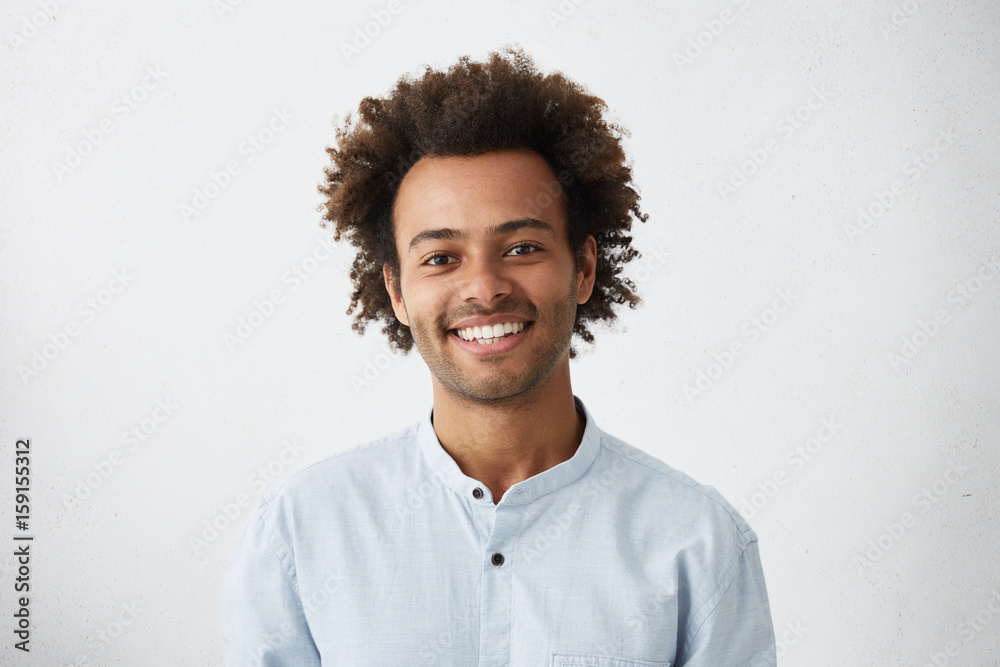 Obraz premium Cheerful African guy with narrow dark eyes and fluffy hair dressed in elegant white shirt having broad smile and good mood while posing in studio over white background. Facial expressions concept