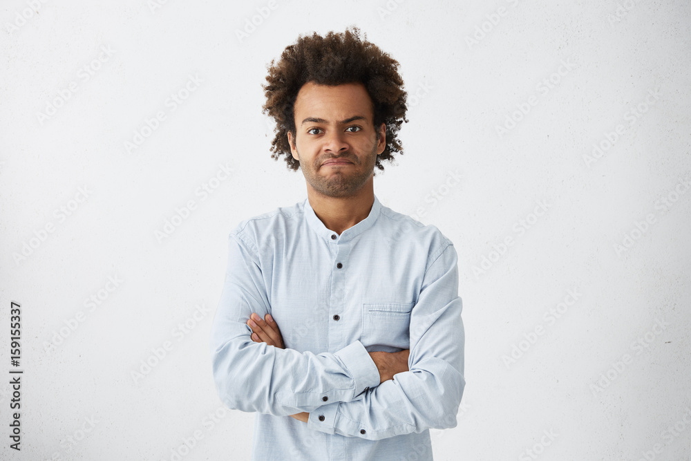 Angry Afro American male having displeased look standing crossed hands against white background. Young freelancer in formal shirt having some problems looking with anger and discontent into camera Angry Afro American male having displeased look standing crossed hands against white background. Young freelancer in formal shirt having some problems looking with anger and discontent into camera
