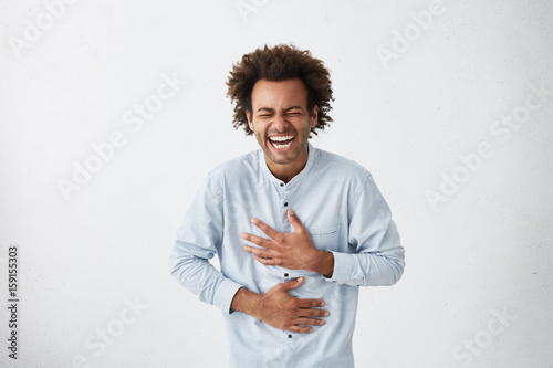 Positive and joyful Afro American male with fine crop of hair bursting into laughing holding his hands on stomach can`t stopping laughing after hearing funny anecdote. Positive emotions and humour