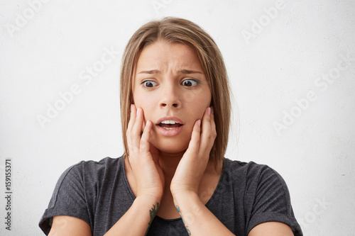 Close-up portrait of puzzled woman with dark bugged eyes holding hands on cheeks looking desperately down. Shocked female having troubles not knowing how to solve them. Emotions and people concept