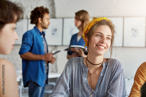 Smiling business lady wearing trendy shirt and yellow head bandage having pleasant smile while having business meeting with her partners isolated over working space. Communication, work concept