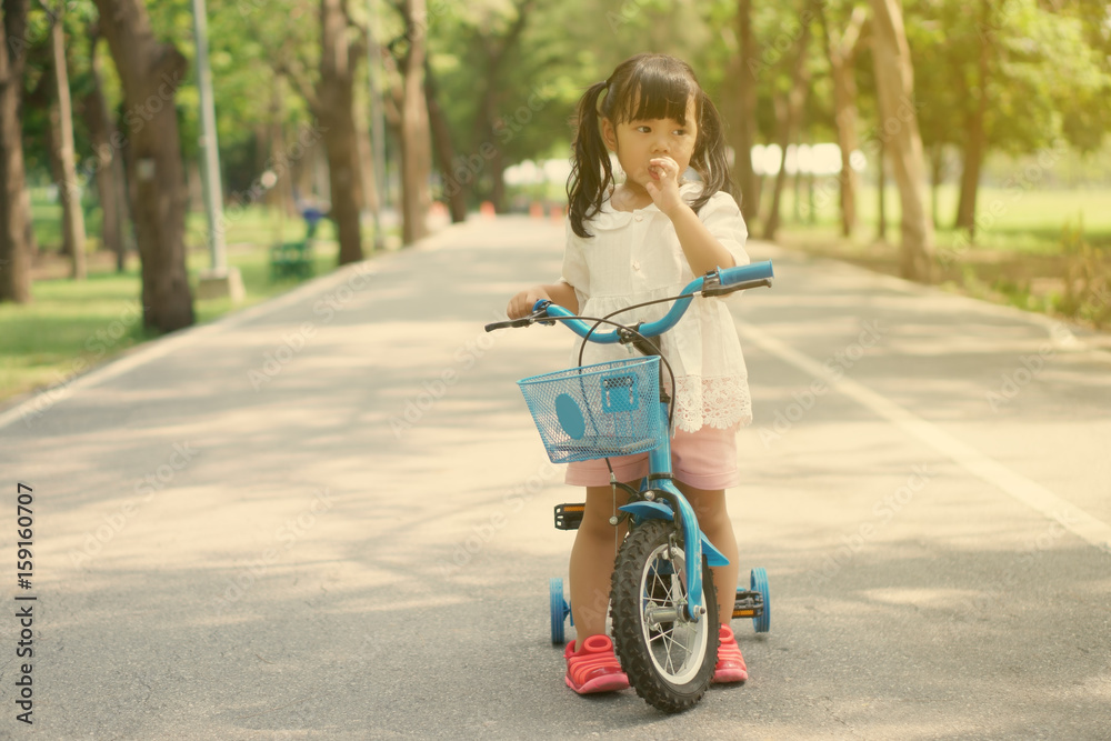 Asian children cute or kid girl cycling on blue bike or bicycle and nail biting on the road at wachirabenchathat public park with green tree for enjoying and exercise on summer holiday with sunlight Asian children cute or kid girl cycling on blue bike or bicycle and nail biting on the road at wachirabenchathat public park with green tree for enjoying and exercise on summer holiday with sunlight