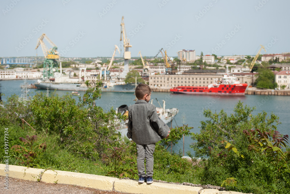 little boy in a jacket stands against the background of a seaport
