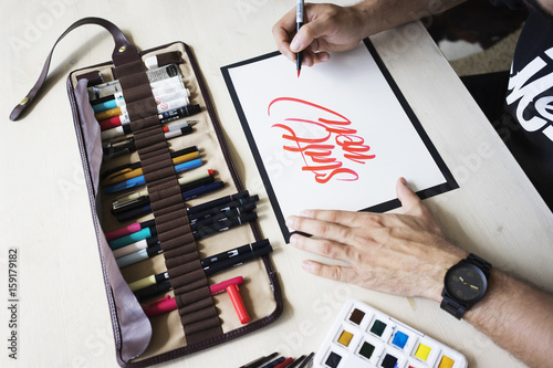 Canvas Print Top close up view on calligrapher who writes phrase Start now on white blank paper with red marker on white wooden table background with markers in leather case and watercolor palette