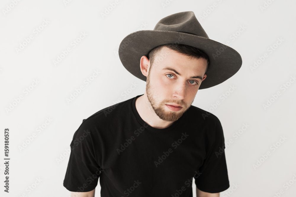 Close up portrait of young bearded guy with blond hair and in grey hat and black t-shirt looks in camera quietly isolated on white background