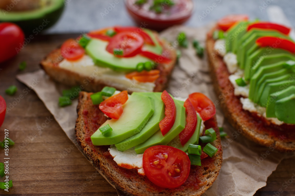 Organic homemade avocado toasts on rye bread with tomatoes, scallions, pepper and cream cheese on vintage wooden board, selective focus. Healthy eating concept