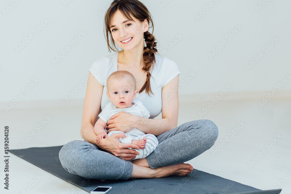 Sport, motherhood and active lifestyle concept - Happy mother and little baby boy sitting on yoga mat and get ready to start workout at home over light background. Sport, motherhood and active lifestyle concept - Happy mother and little baby boy sitting on yoga mat and get ready to start workout at home over light background.