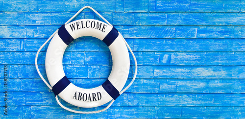 White color Life buoyancy with welcome aboard on it hanging on blue wall. had space on right side for your text.