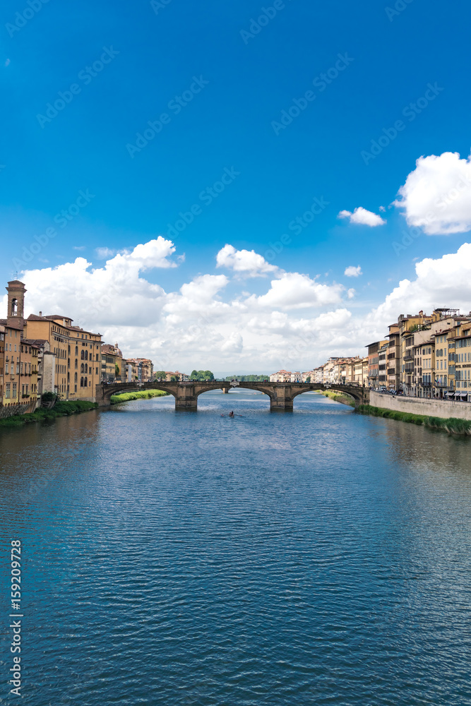 Vertical composition the River Arno in Florence with puffy clouds and rowers on the water.
