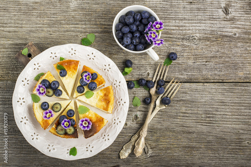 Homemade fresh cheesecake with blueberries and edible flowers on a white ceramic plate, on a simple wooden background. From the top view. The concept is helpful comfortable food