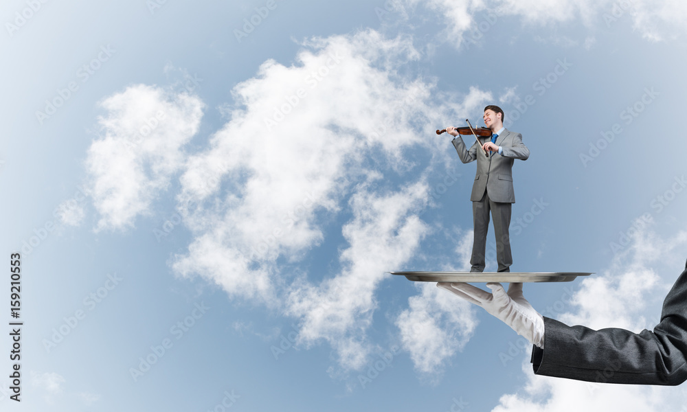 Businessman on metal tray playing violin against blue sky background Businessman on metal tray playing violin against blue sky background