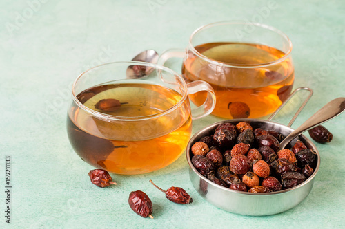  Rose hips, dried fruit in a metal bowl and freshly prepared tea in clear glass mugs on a light green background.
