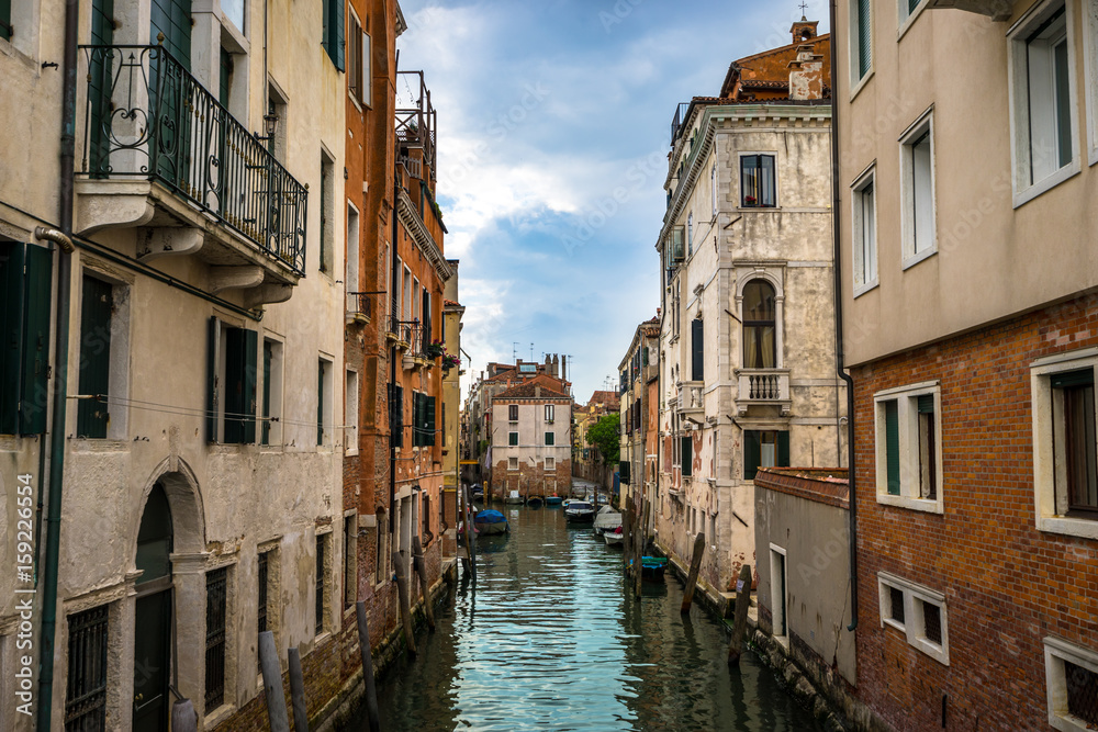 Panoramic view on famous Grand Canal among historic houses in Venice, Italy at cloudy day with dramatic sky, wood bridge and sitting woman on the foreground.