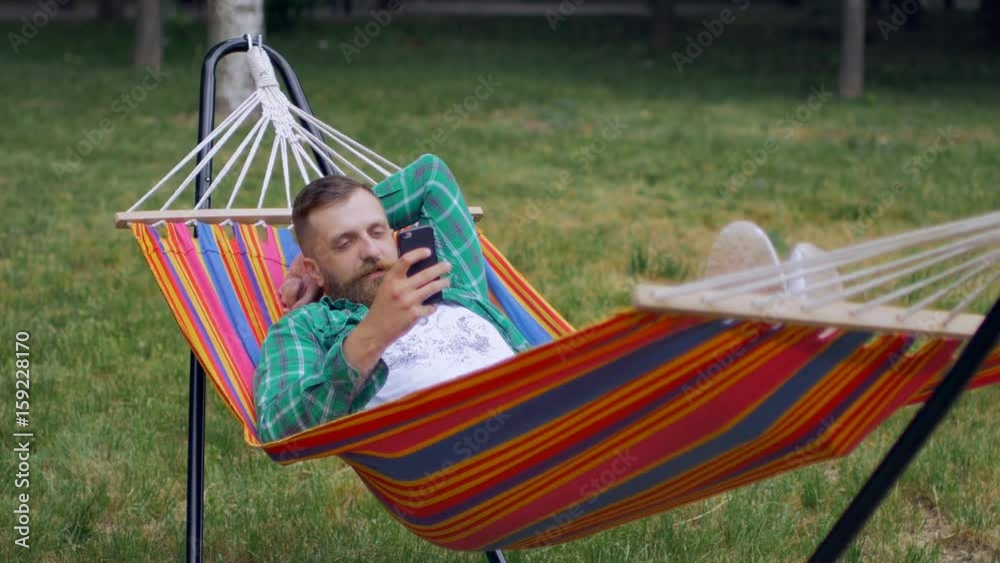 A bearded man in a green checkered shirt lies on a hammock and uses a telephone A bearded man in a green checkered shirt lies on a hammock and uses a telephone