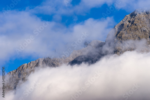 Fiordland National Park, New Zealand - March 16, 2017: Tall mountain peaks, lining Milford Sound, pit their heads through white clouds under blue sky.