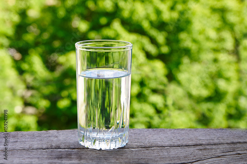 Glass with water and mint on a wooden table against a background of green foliage